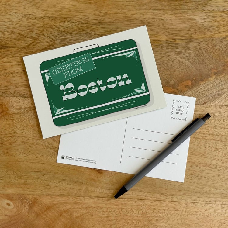 The front and back sides of a postcard with a pen placed on top. The front features a vintage suitcase illustration and the back shows the address lines and stamp indicator.