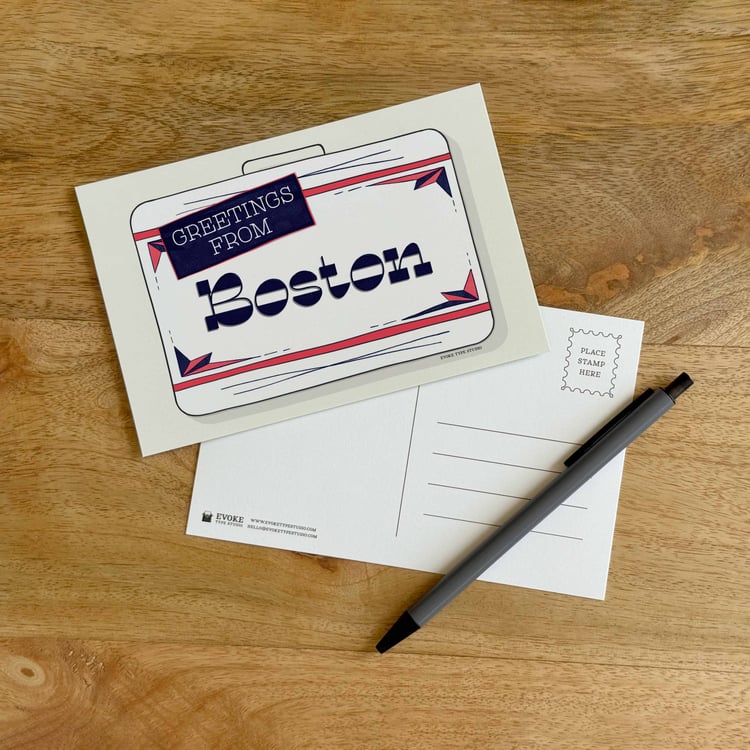 The front and back sides of a postcard with a pen placed on top. The front features a vintage suitcase illustration and the back shows the address lines and stamp indicator.