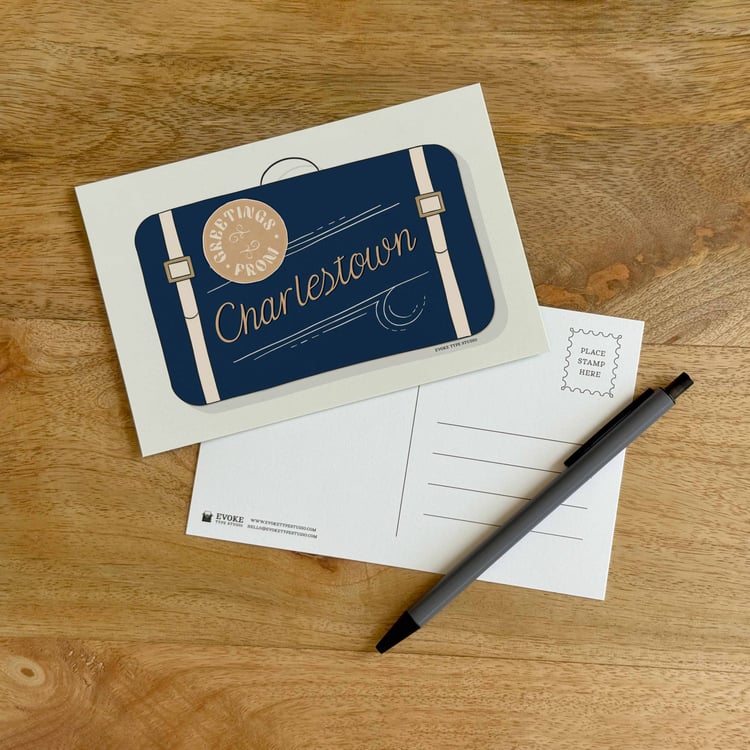 The front and back sides of a postcard with a pen placed on top. The front features a vintage suitcase illustration and the back shows the address lines and stamp indicator.