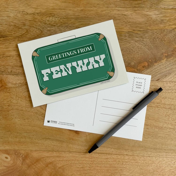 The front and back sides of a postcard with a pen placed on top. The front features a vintage suitcase illustration and the back shows the address lines and stamp indicator.