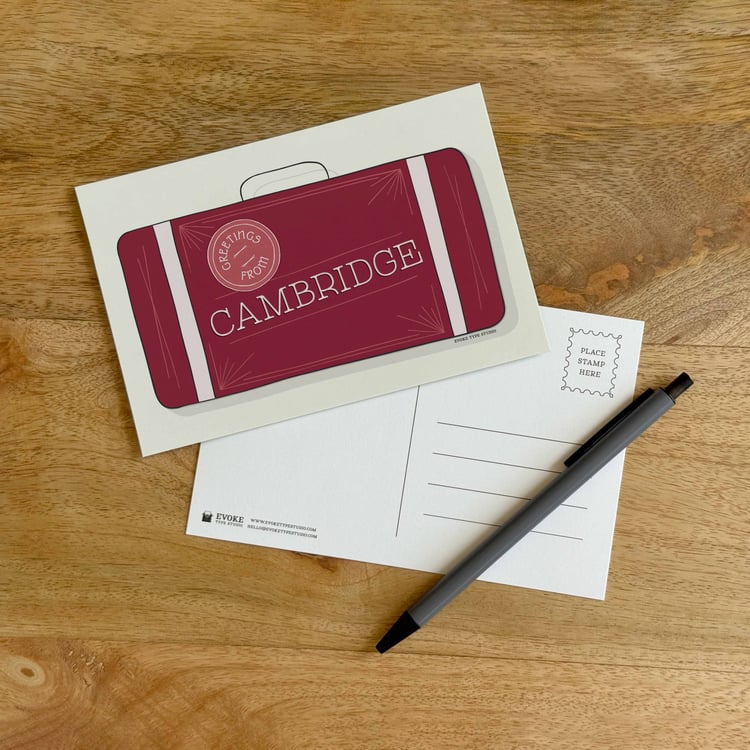 The front and back sides of a postcard with a pen placed on top. The front features a vintage suitcase illustration and the back shows the address lines and stamp indicator.