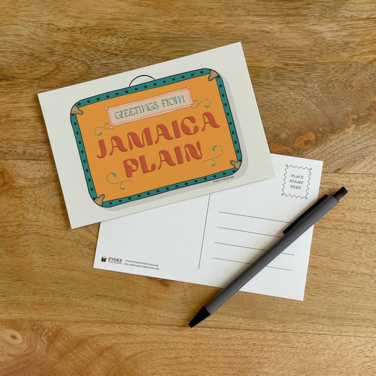 The front and back sides of a postcard with a pen placed on top. The front features a vintage suitcase illustration and the back shows the address lines and stamp indicator.