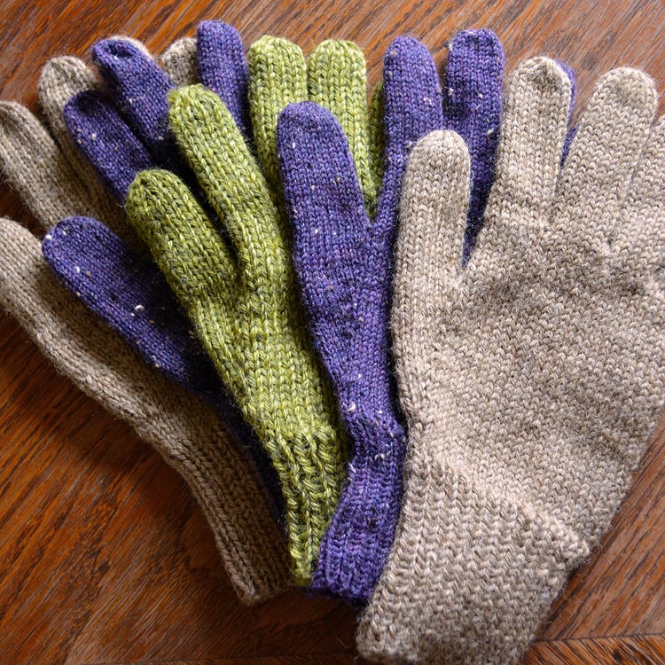 A spread of knit gloves of various sizes, yarn weights, and colours on a wooden tabletop. Gloves have ribbed cuffs and plain stockinette bodies, and are knit in bulky, worsted, and fingering weight.