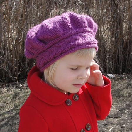 A young blond girl in a red coat wears a purple chunky-knit slouchy cap with a staghorn-cabled hatband.