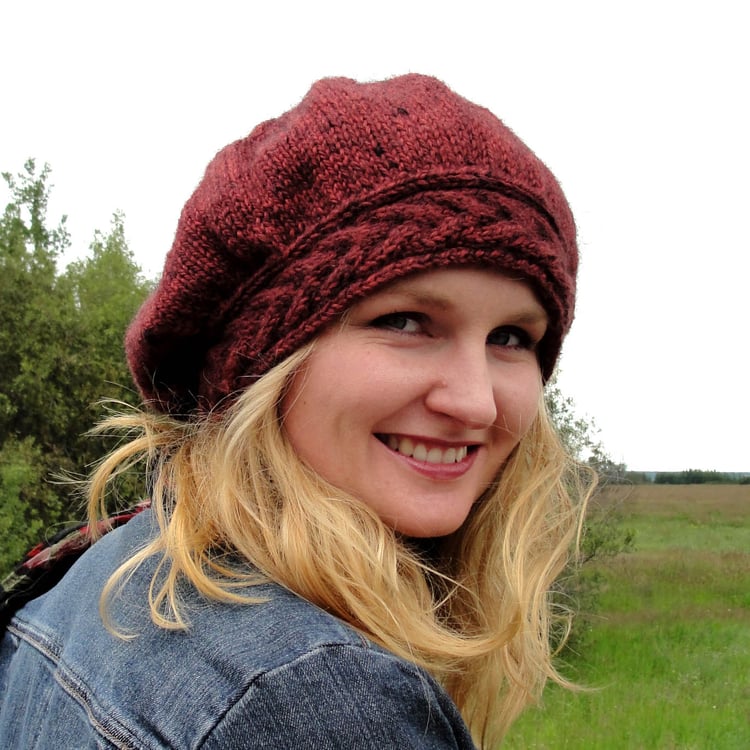 A blond Caucasian woman smiles at the camera and wears a burgundy chunky-knit slouchy hat with a staghorn cabled hatband.