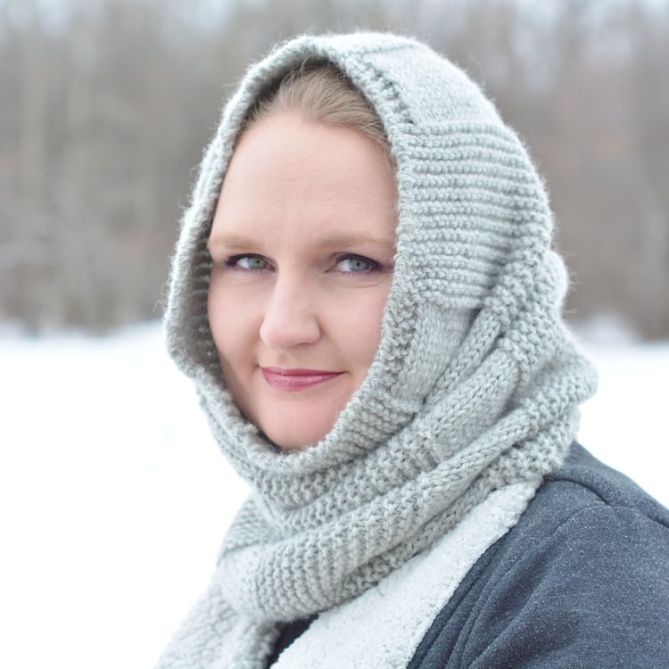 Talena, a blond Caucasian woman, wears a tubular cowl around her face like a hood. The cowl has alternating blocks of garter and stockinette stitch.