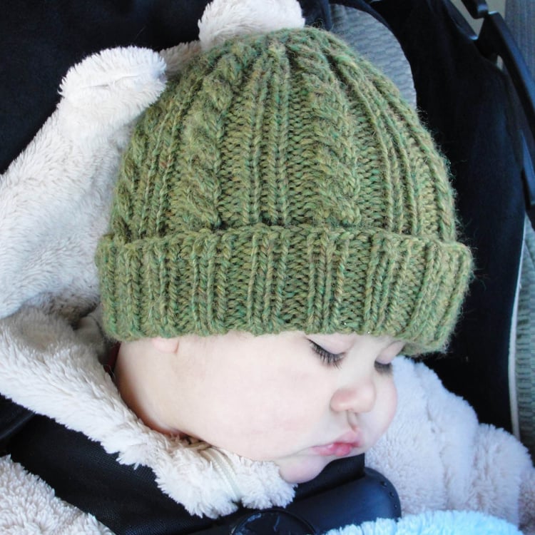 A sleeping baby boy in a car seat wears a green cabled beanie alternating with a single rib in the repeat with a ribbed rolled brim.