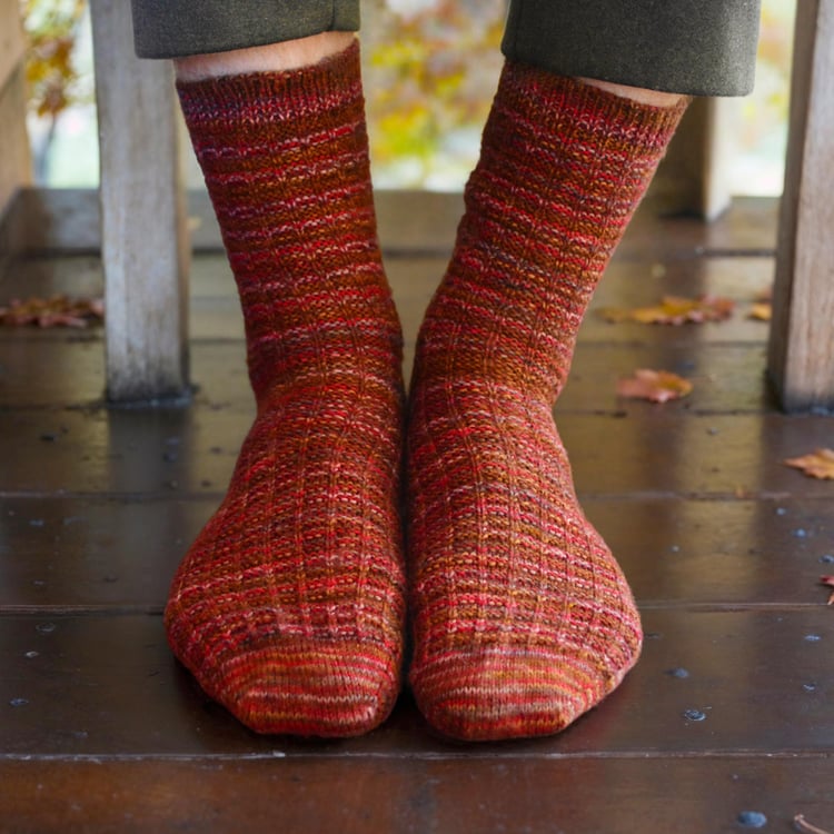 Closeup of toes and legs of Adventure Socks knitting pattern on models feet among autumn leaves on a rustic deck, showing the raglan toe shaping the waffle weave texture.