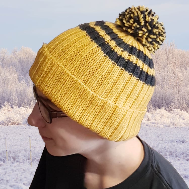 Teenage boy in profile wears a yellow ribbed toque with two black stripes, a two-colour pompom, and a rolled brim in front of a snowy landscape.