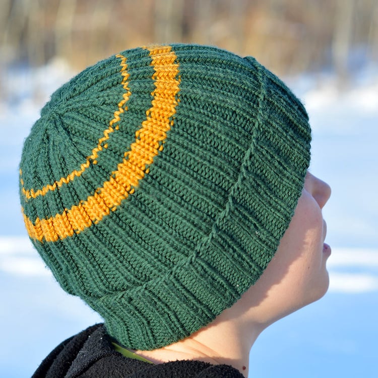 A teenage boy looks away at the snow and wears a jade green rolled-brim ribbed knit watchmans beanie with two irregular golden stripes near the crown, which is shaped using raglan decreases.
