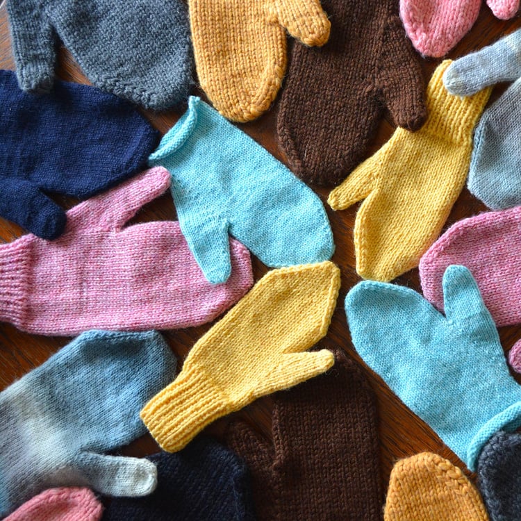 A flat lay of a pinwheel of knit mittens of various colours, yarn weights, and sizes. All have solid colours, stockinette bodies, thumb gussets, and tops tapered at angles to a blunt end. The cuffs are varied lengths of one-by-one ribbing, with a few fing