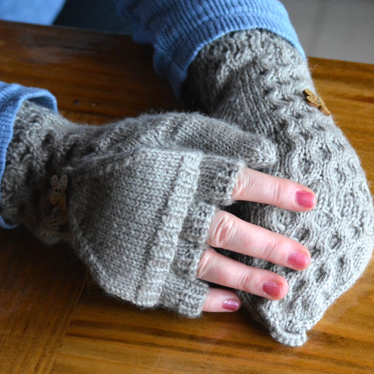 Womans hands on table wearing knit fingerless flip-top gloves (glittens) with all-over honeycomb cables. The flip-top is held with a wooden butterfly button secured through a crochet loop on the end.