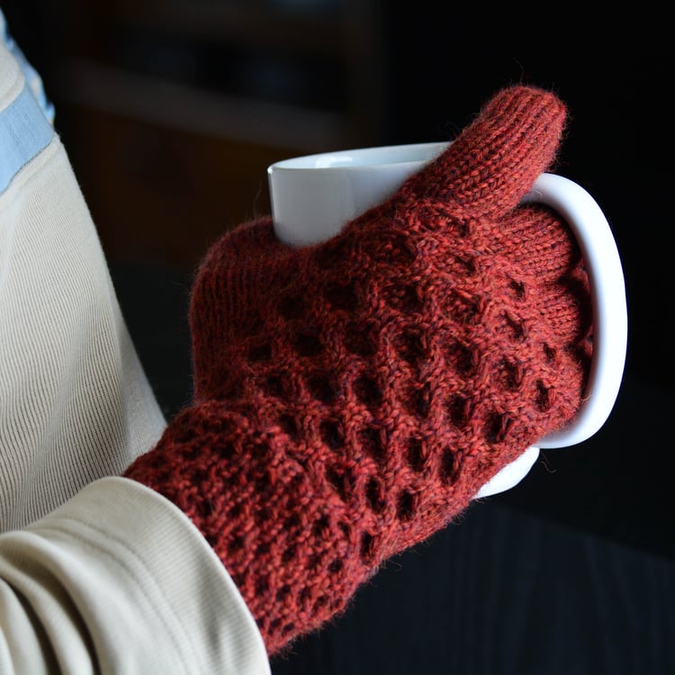 Closeup of a mans hand wearing a size 8 The Honey Tree knit glove in Rooibos red with all-over 4-stitch honeycomb cables on the back and 2-stitch honeycomb cables on the long cuff. He holds a mug.