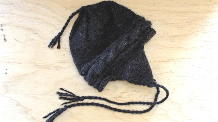 Flat lay of a knit chullo hat with earflaps and twisted yarn ties with tassels at the end, a twisted yarn tie at the top of the crown, and a twisted rope cable hatband.