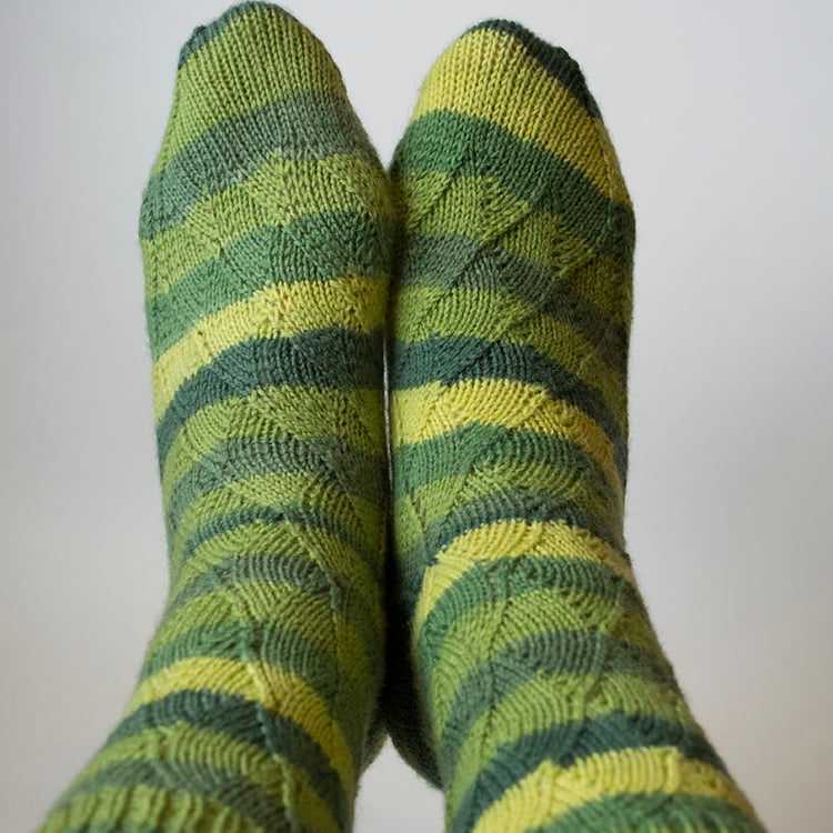 Feet held up displaying a pair of Beanstalk Socks, no-cable variant. Pretty nested scallops climb the leg in a repeating pattern to a one-by-one ribbed cuff in mirror symmetry on each foot.