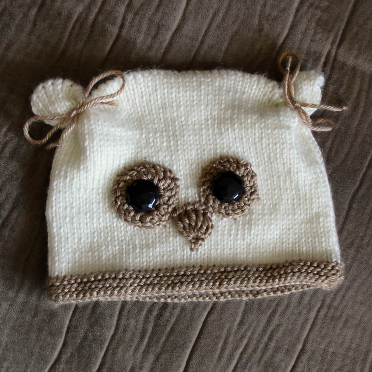 A rectangular cream-coloured knit childs hat lays on a couch. It has yarn bows around the top corners to make ears, and an owl face made with beige crochet applique features and black buttons.
