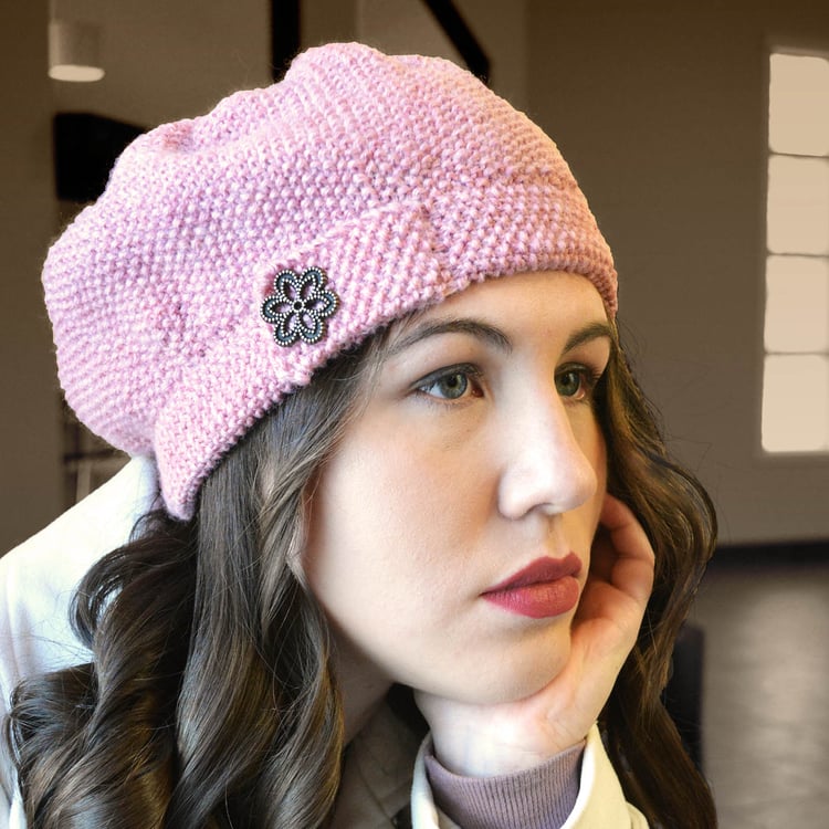 Wistful young woman wears a chic heather pink knit beret with all-over seed stitch, a gentle flare to the crown, and a hatband with a button tab and large brushed brass flower button.