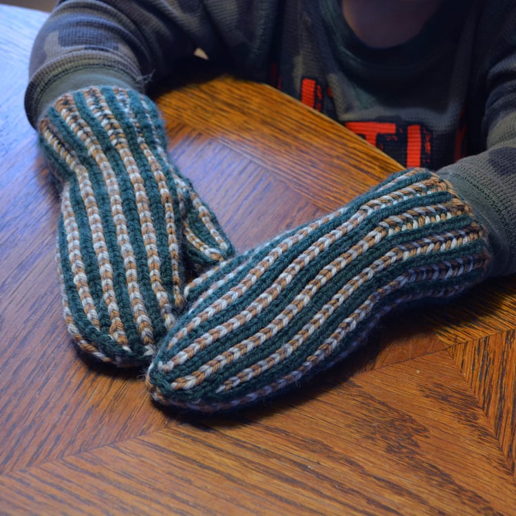 Close-up of a little boys hands wearing striped three-colour brioche-knit Revolution Mittens, with stripes of green alternating with stripes of alternating beige and grey.