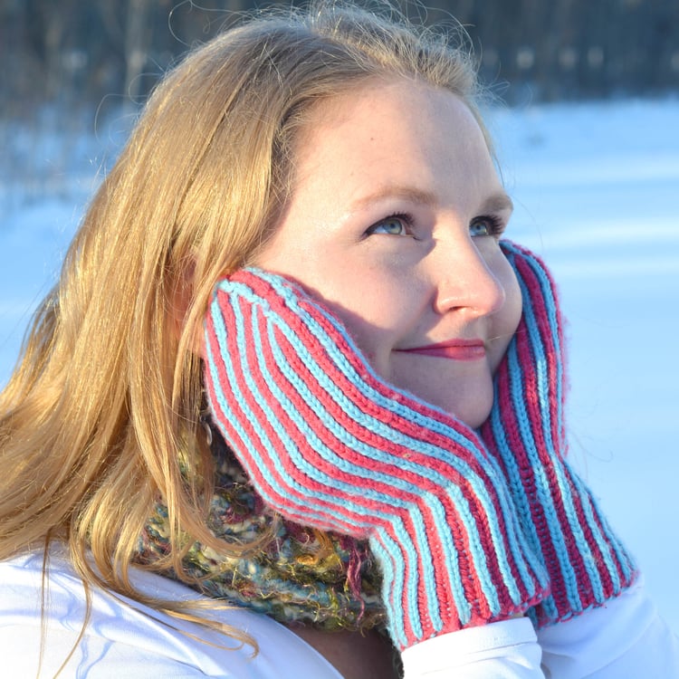 Blond woman looks sweet and holds hands to her face in front of the snow while wearing candy-pink and sky-blue striped brioche-knit mittens with a long cuff.