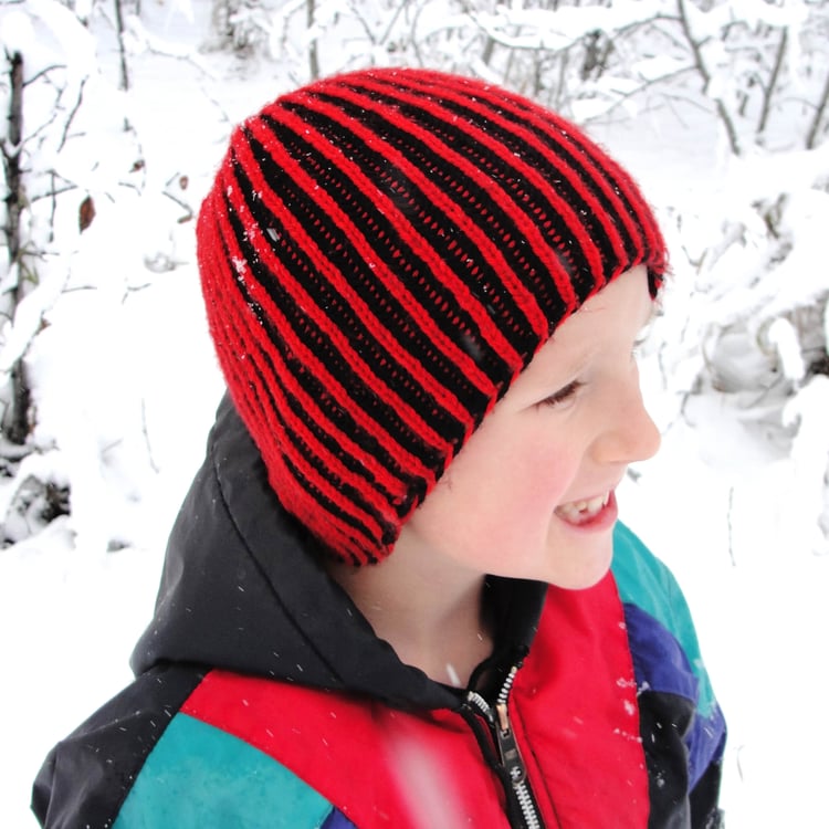 Little boy wears a brimless brioche-knit beanie that covers his ears. The ribs have red on the top and black behind. Hes standing in front of a snowy background.