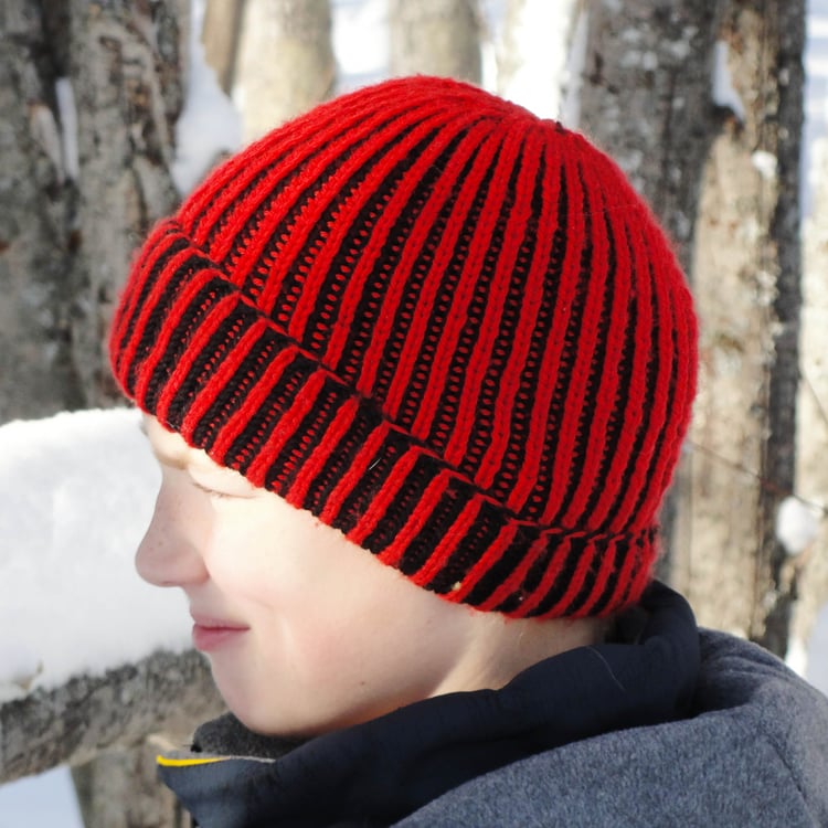 Pre-teen boy in profile wears a seamless two-colour brioche-knit beanie with a rolled brim that has red-and-black vertical stripes along the ribs in front of a snowy treed background.