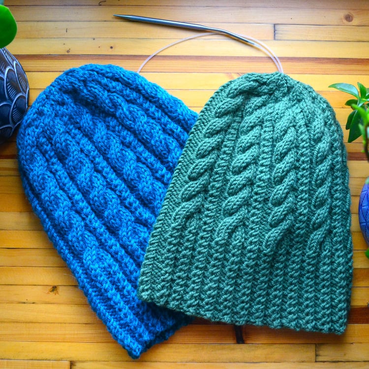 A dark green cable-knit toque lays at an angle on a matching turquoise beanie on a golden wooden surface between two small potted plants. A circular needle is tucked beneath them.