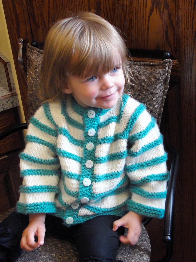 Toddler girl sitting in a chair and wearing a turquoise and white long-sleeved seamless chunky-knit Asian-inspired baby cardigan with textural stripes, raglans shoulders, stand-up collar, and a button band.