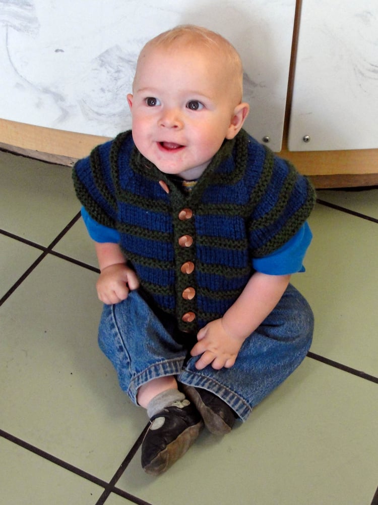 Baby boy sitting up while wearing a sleeveless, seamless chunky-knit Asian-inspired baby cardigan with textural stripes, raglans shoulders, stand-up collar, and a button band.