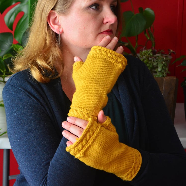 Woman holding hands next to face and looking sideways while wearing golden fingerless mittens with a twisted cable at the top and bottom cuffs.
