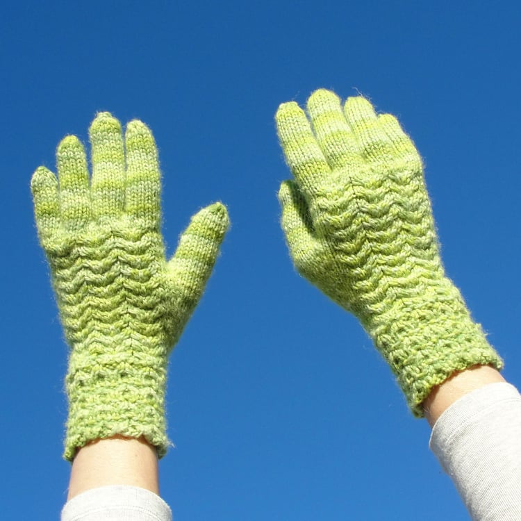 Hands reach up for the blue sky wearing light green knit gloves with scale-like 4-stitch cables on the back and 2-stitch cables on the long cuff.