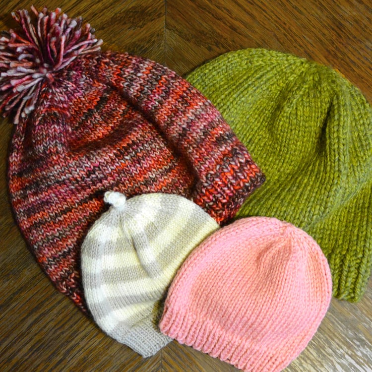 Rounded knit beanies of various sizes, yarn weights, and colours overlap on a table. All have ribbed unrolled brims and rounded stockinette crowns. Toppers: pompom, tied I-cord knot, and none.