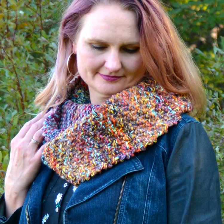 Woman with purple and blond hair wears a multicoloured textured chunky knit cowl with colour gradient over a denim blazer and looks downward.