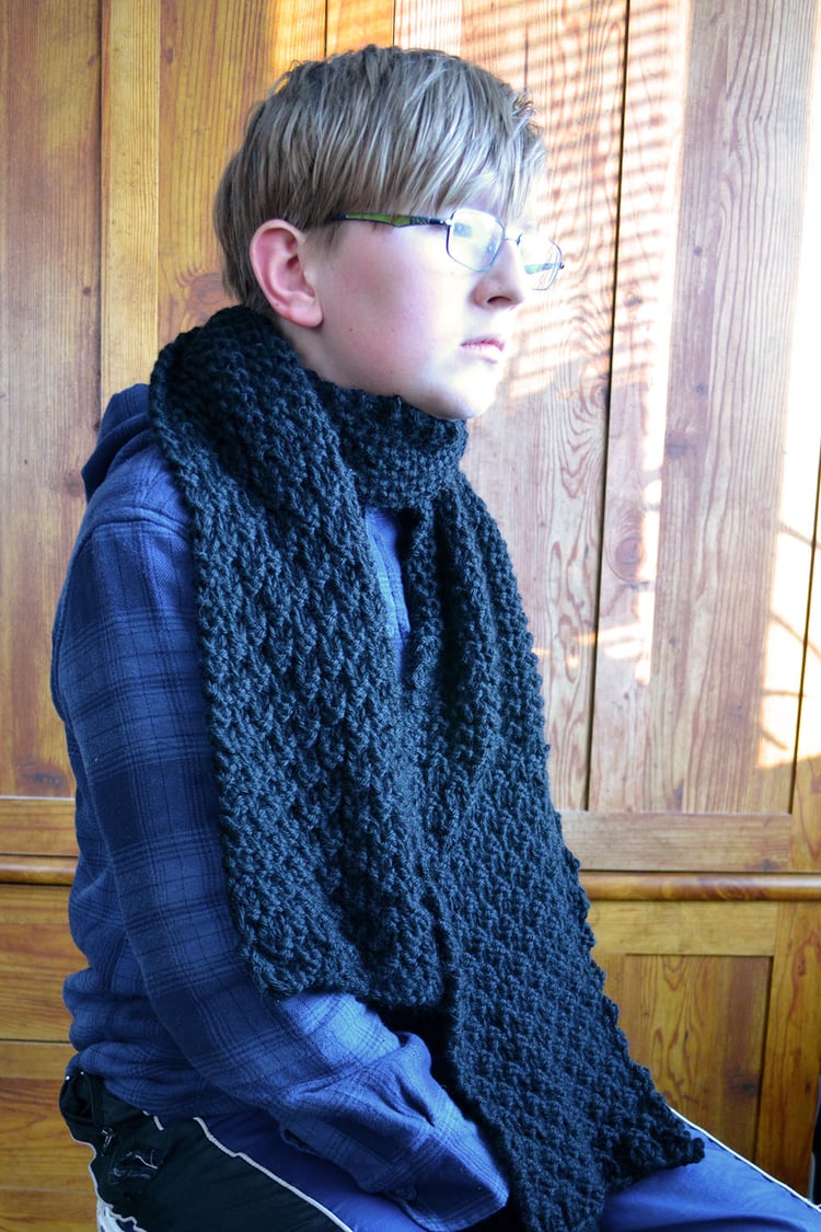 Young teenage boy wears a black textured knit scarf with stitch patterns in different scales and looks off-camera while sitting.