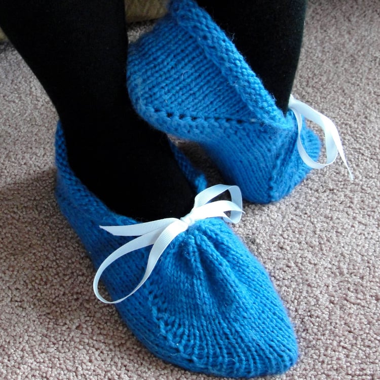 Womans feet wearing pretty blue knit slippers in chunky yarn. The ankle rolls down, and the front is gathered by a ribbon tied with a bow. Short row shaping in front and back.