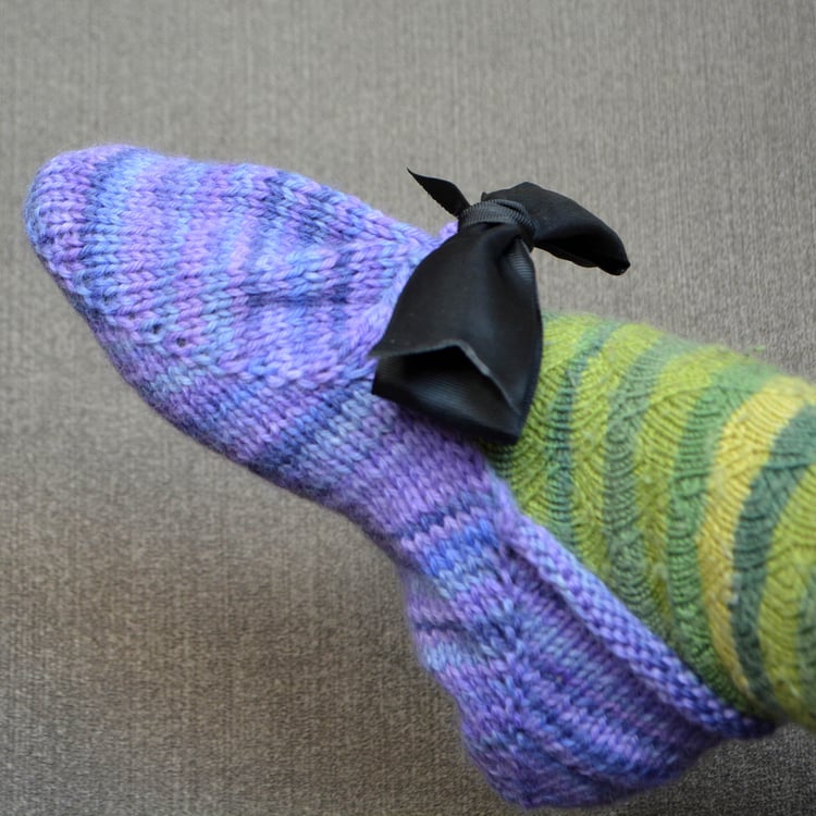Womans foot wearing pretty variegated purple knit slippers in worsted yarn. The ankle rolls down, and the front is gathered by a ribbon tied with a bow. Short row shaping in front and back.
