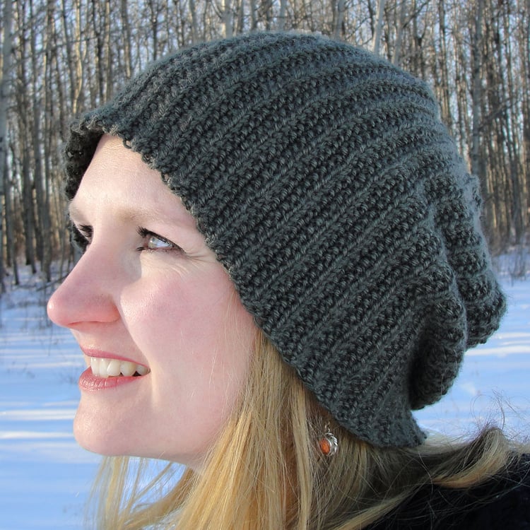 Woman wearing a grey slouchy snowboarder knit beanie made with broken rib in front of snowy trees.