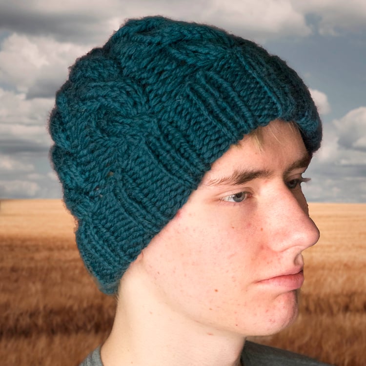 Young Caucasian man wears a dark teal blue chunky-knit cabled cap with a rolled brim and looks off-camera while standing in front of a golden wheat field and a stormy sky.