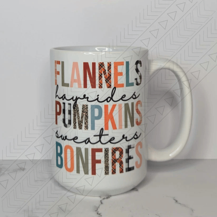 A white ceramic mug with the words "FLANNELS," "HAYRIDES," "PUMPKINS," "SWEATERS," and "BONFIRES" stacked vertically. The text is in a mix of different fonts and colors, including solid orange, green, and brown, as well as plaid and leopard print patterns