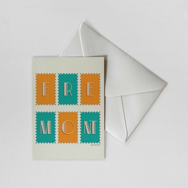 A notecard of postage stamps featuring the Fremont neighborhood in Seattle, Washington drawn in a unique lettering style with a white envelope. Artwork has a blue and orange color scheme