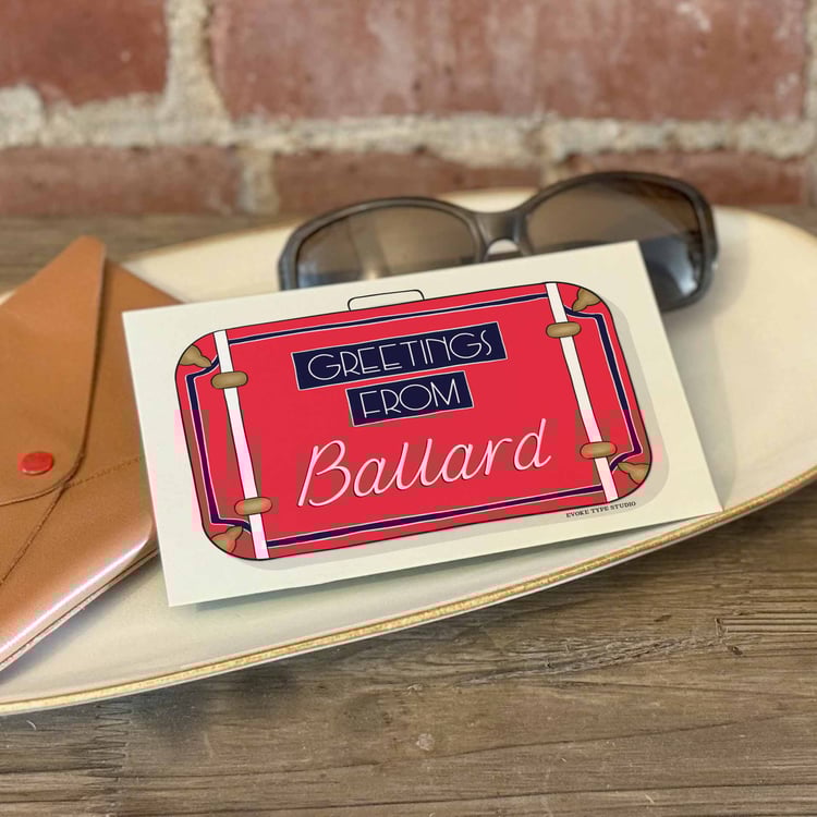 The front and back sides of a postcard with a pen placed on top. The front features a vintage suitcase illustration and the back shows the address lines and stamp indicator.