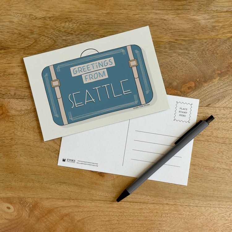 The front and back sides of a postcard with a pen placed on top. The front features a vintage suitcase illustration and the back shows the address lines and stamp indicator.