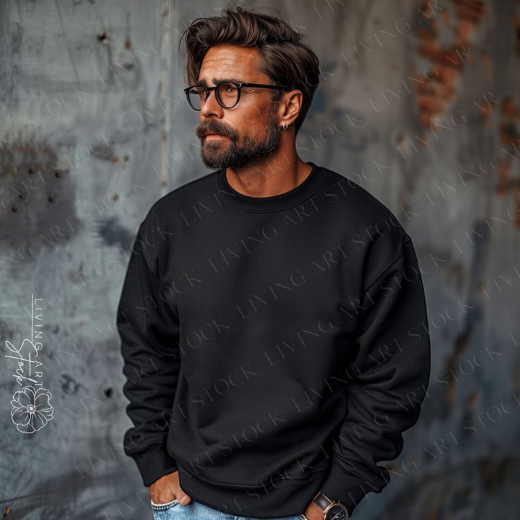 A ruggedly handsome man with a full beard, dark hair, and glasses stands against a textured, distressed metal background, modeling a plain, black crewneck sweatshirt. He is looking off to the side with a serious, thoughtful expression, his hands casually 