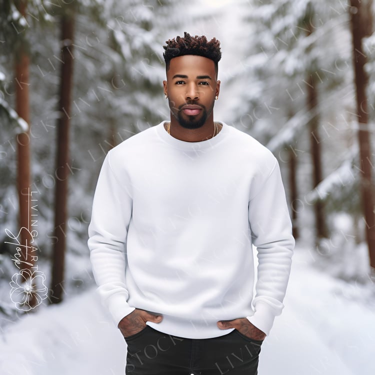 Black man wearing white crewneck sweatshirt standing with his hands in his pockets, he is standing in a snow covered forest with good natural light