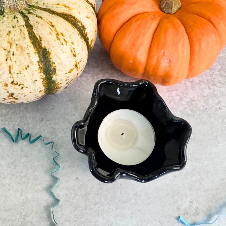 A top view of a black tea light candle on a grey table with pumpkins and blue crinkled paper props.