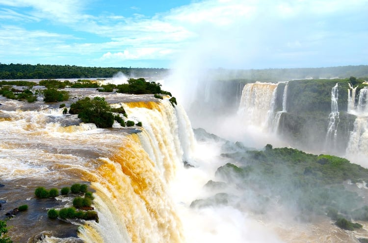 Top-down drone view of Iguaçu Falls releasing massive turquoise plumes with a bright rainbow arc spanning the mist.