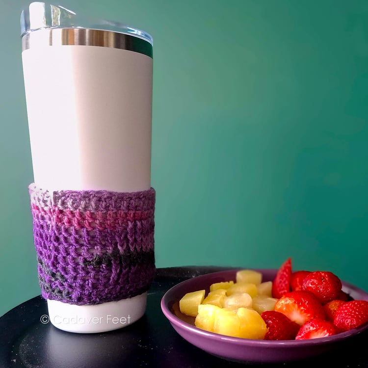 a purple crochet mug hugger on a white tumbler next to a fruit plate