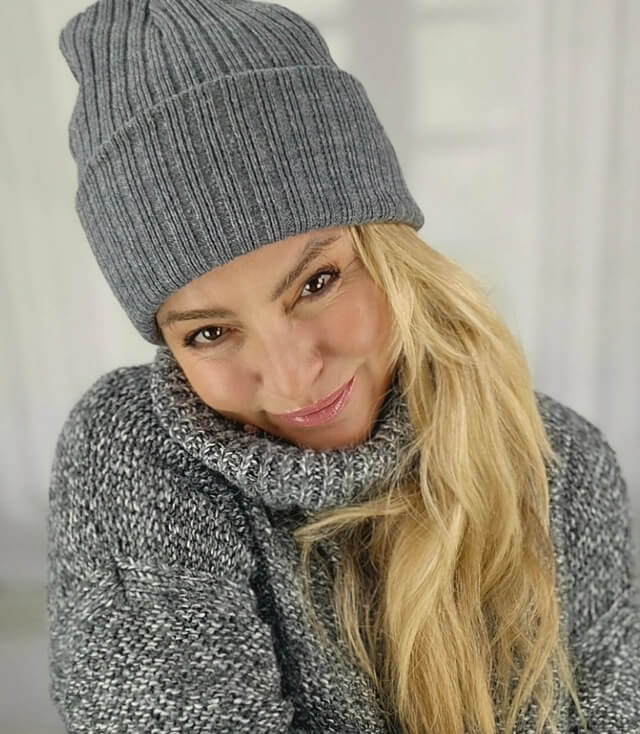 Frau mit langem, blonden Haar, die in einem grauen Pullover und einer grauen Strickmütze lächelt.