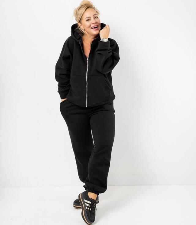 Frau in schwarzem Hoodie und Jogginghose, lächelt und posiert vor weißem Hintergrund.