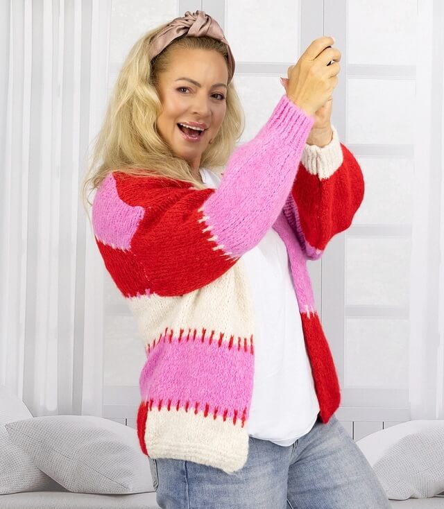 Frau mit blonden Haaren trägt einen bunten Cardigan in Rot, Pink und Creme, lächelnd vor einer hellen Wand.