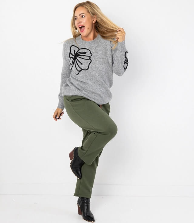 Dame in grauem Pullover mit schwarzem Blumenmuster und olivgrüner Hose, fröhlich posierend vor weißem Hintergrund.